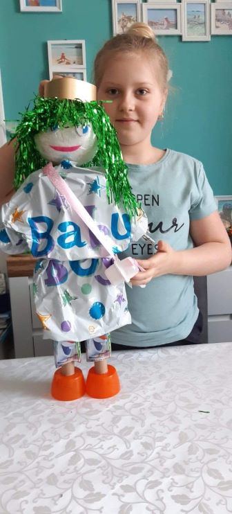 grafika przedstawia dziewczynk z wykonan lalk. Lalka ma na gowie ma burze lokw w kolorze zielonym. Ubrana jest w sukienk z plastikowej foli z nadrukiem, przepasana jest torebk wykonan z wstki i kubeczkw po serku. Buciki lalki to pomaraczowe nakrtki.